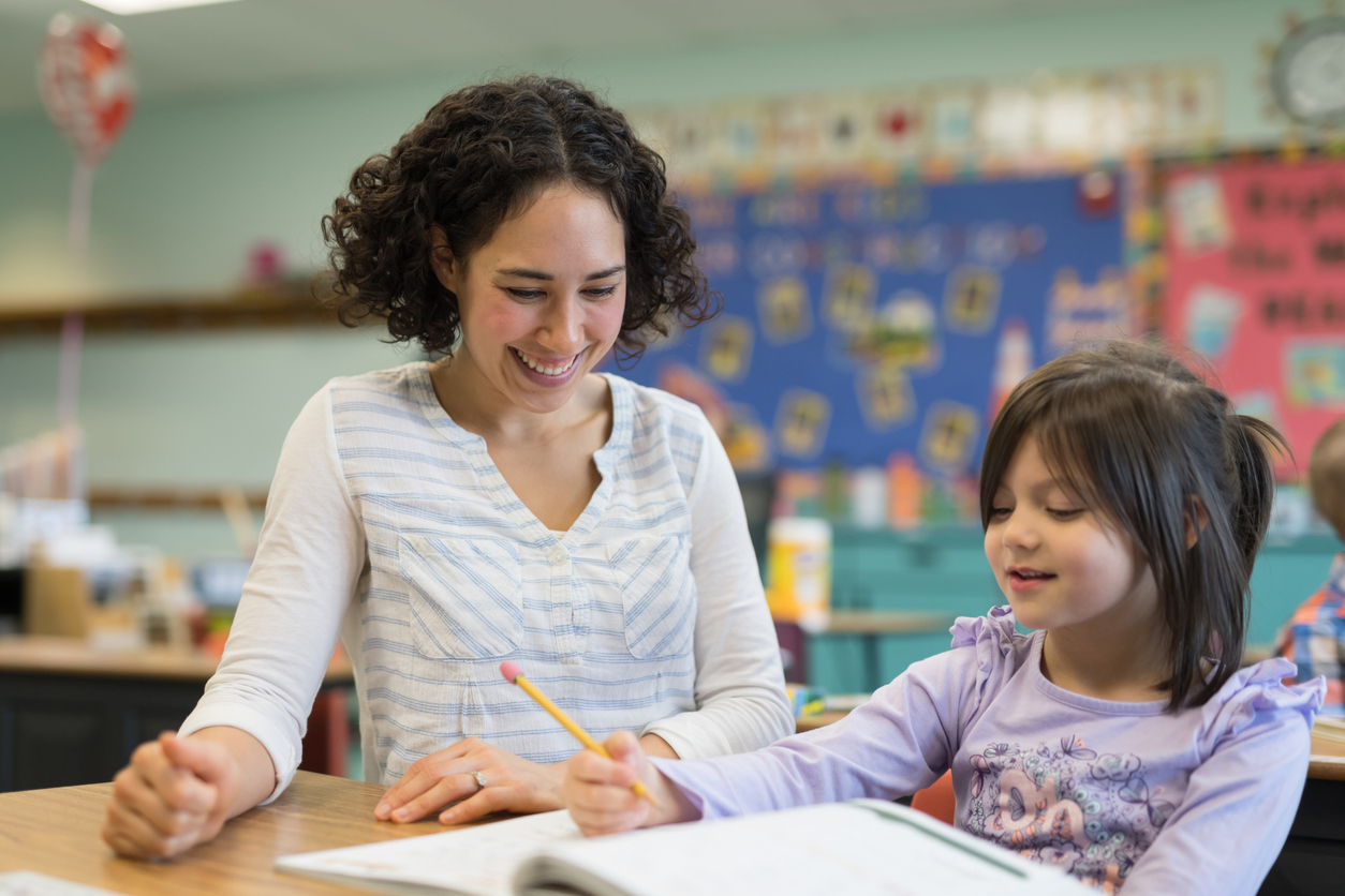 A young girl cheerfully writes in her spelling workbook at her desk. Her teacher is kneeling beside the desk and encouraging her.